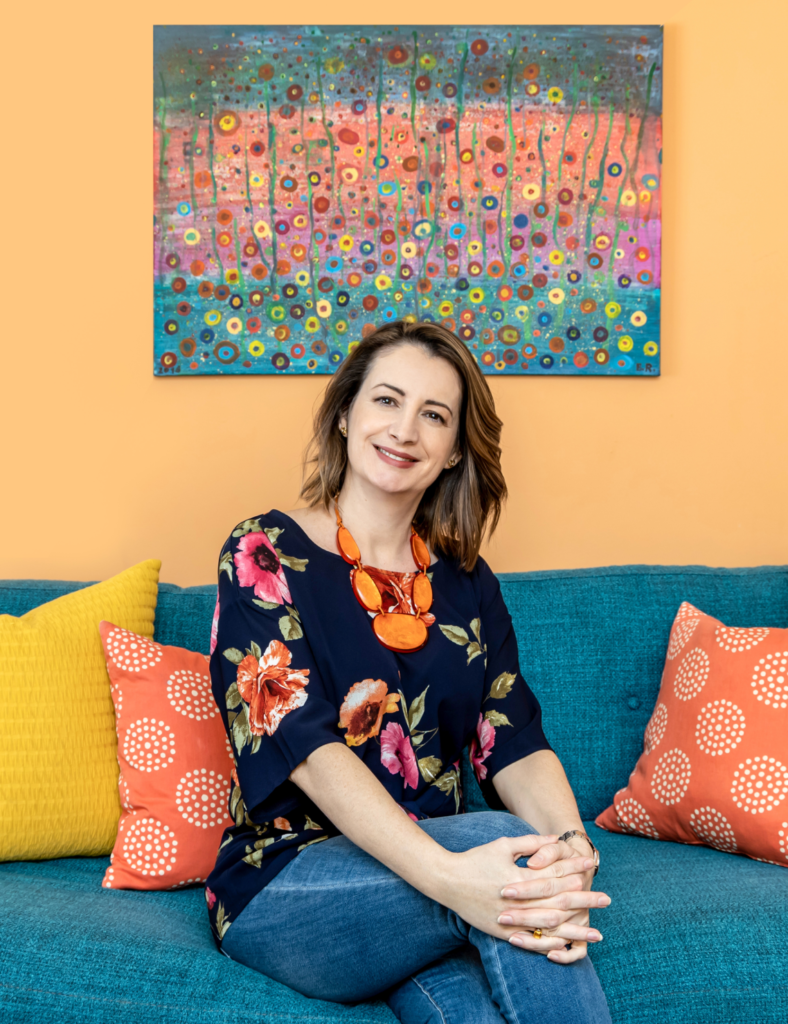 Andrea Adamicka, founder and decorator of Farbaa Interiors sitting on a blue-green couch with red and yellow cushions, and an abstract artwork in the backdrop.