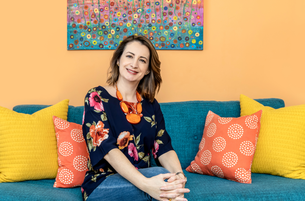 Andrea Adamicka, founder and decorator of Farbaa Interiors sitting on a blue-green couch with red and yellow cushions, and an abstract artwork in the backdrop.